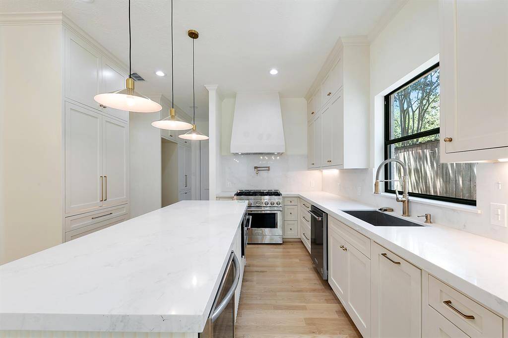 This modern kitchen features sleek white cabinetry, a large island with pendant lighting, stainless steel appliances, and a spacious layout. Natural light pours in through a sizable window, highlighting the elegant finishes and ample counter space.