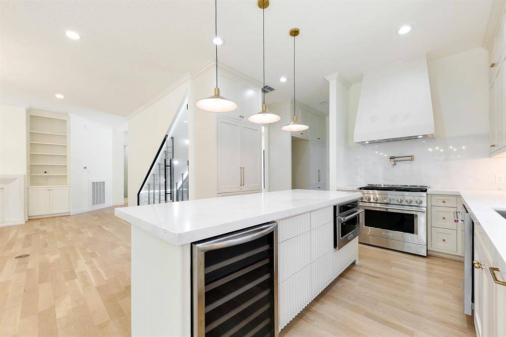 This photo showcases a modern kitchen with sleek white cabinetry, a large island with a wine cooler, and elegant pendant lighting. It features a high-end gas range and hood, bright hardwood floors, and an open layout leading to a living area with built-in shelving.