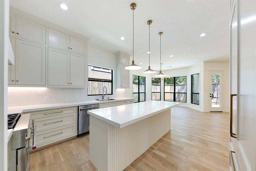 This modern kitchen features an open layout with hardwood floors, a large central island with a sleek white countertop, and stylish pendant lighting. Ample cabinetry provides plenty of storage, and large windows offer abundant natural light, creating a bright and inviting space.