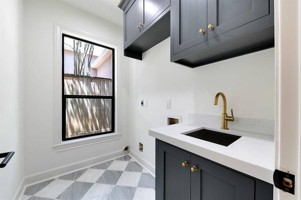 This photo showcases a stylish laundry room with modern gray cabinetry, a sleek gold faucet, and a white countertop. The room features a window for natural light and a chic checkered floor design.