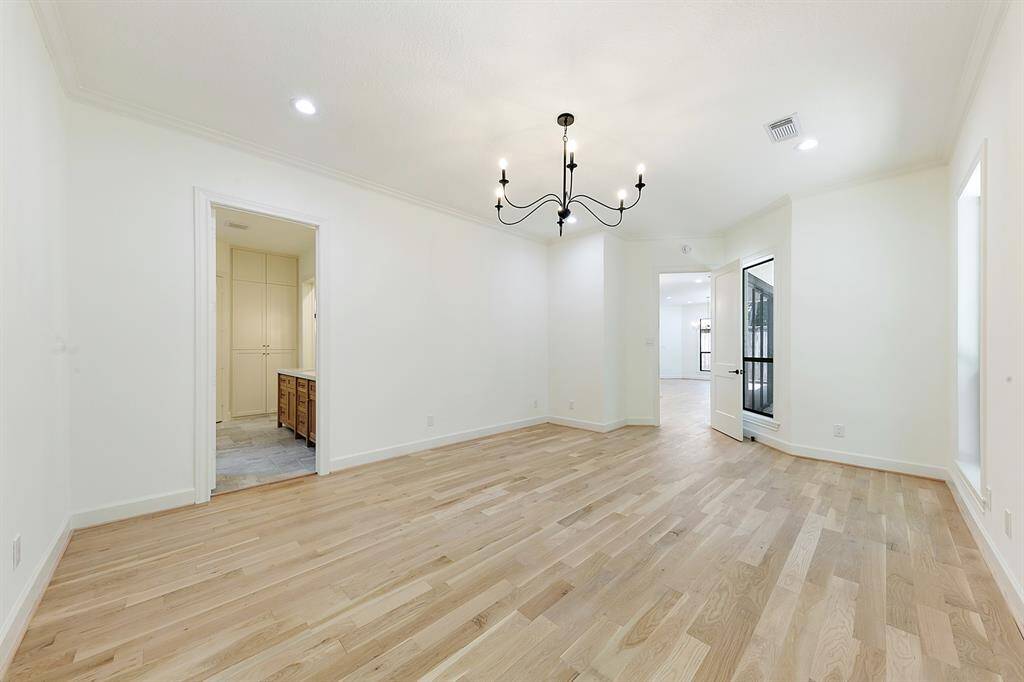 This room features light hardwood floors, a modern black chandelier, and recessed lighting. It connects to a bathroom and a hallway, with large windows providing natural light.