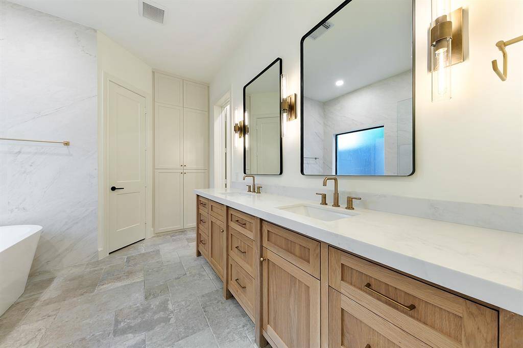 This bathroom features a modern, elegant design with dual sinks, sleek brass fixtures, and wooden cabinetry. It has large mirrors, a spacious layout, and a luxurious soaking tub, creating a serene and upscale atmosphere.