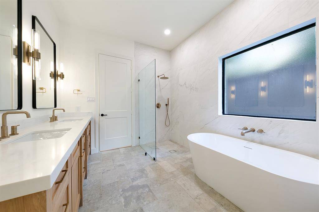 This modern bathroom features a spacious layout with a double vanity, sleek fixtures, a glass-enclosed shower, and a freestanding tub. Large frosted windows provide natural light while maintaining privacy.