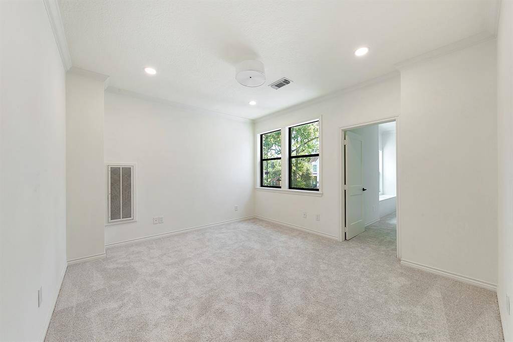 This is a spacious, well-lit bedroom with neutral carpeting and walls. It features large windows for natural light, recessed lighting, and an adjoining room.