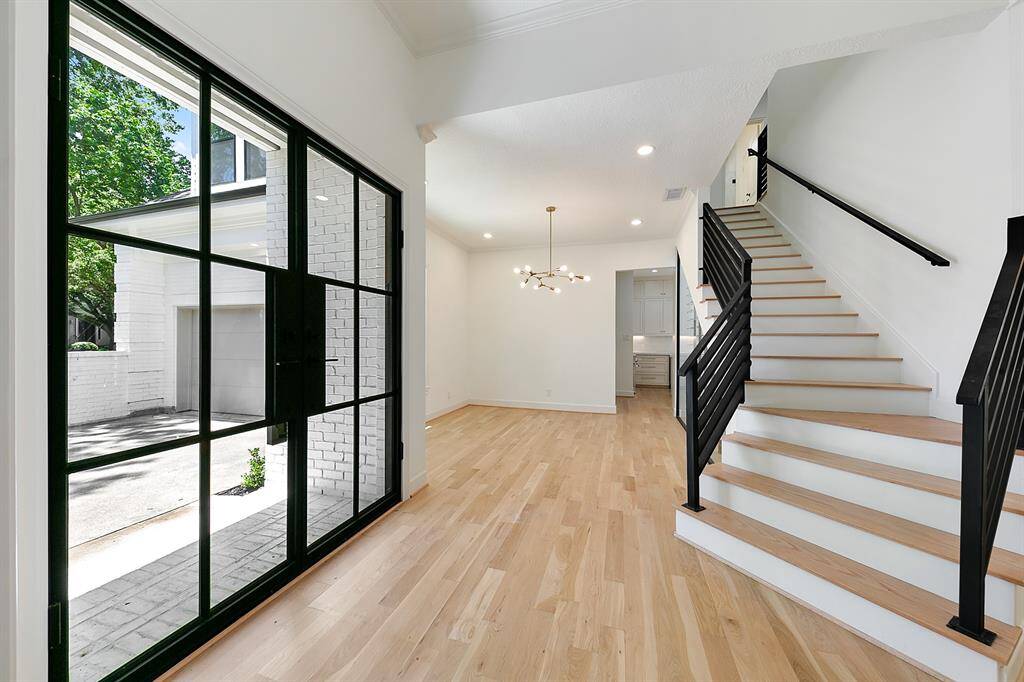 This bright entryway features modern glass doors, light wood flooring, and a stylish staircase with black railings. An elegant light fixture enhances the open, inviting space.