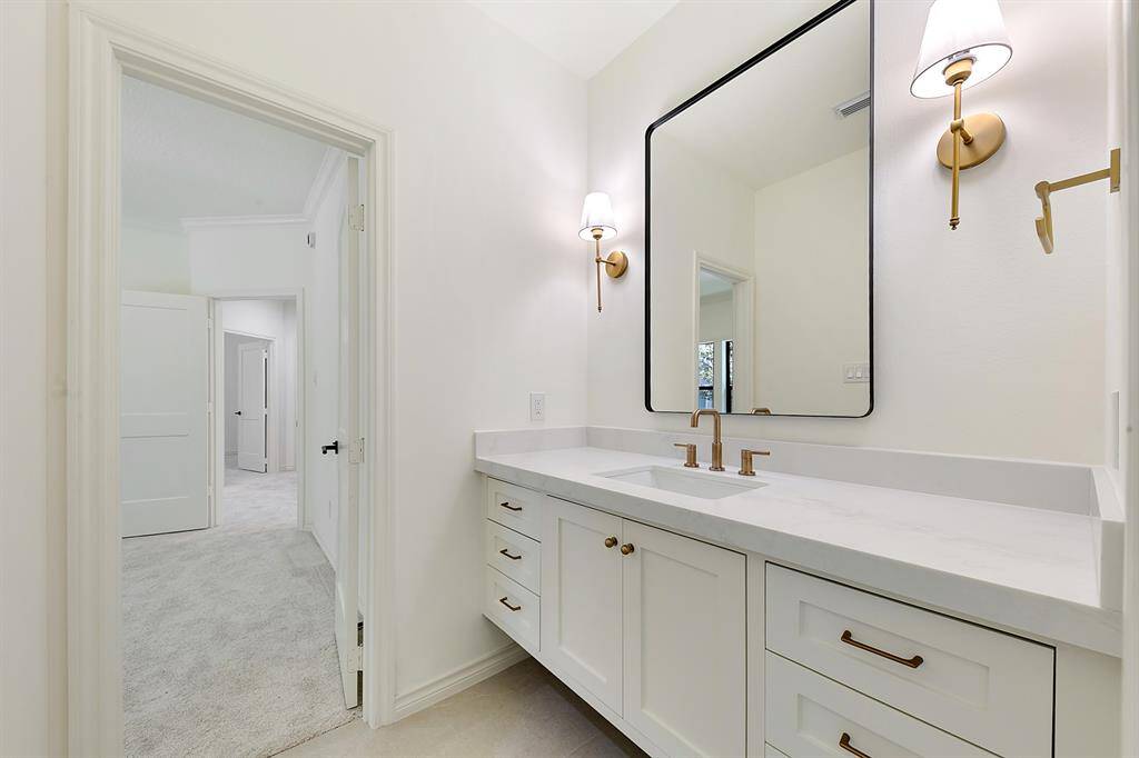 This photo showcases a bright, modern bathroom with a large mirror, elegant lighting, and a sleek vanity with ample storage. The clean design features white cabinetry and gold fixtures, leading into a carpeted hallway.
