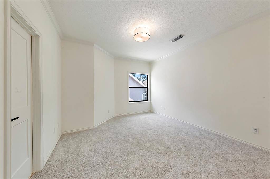 This room is bright and spacious with neutral walls and carpeting, featuring a single window for natural light and a modern ceiling light fixture.