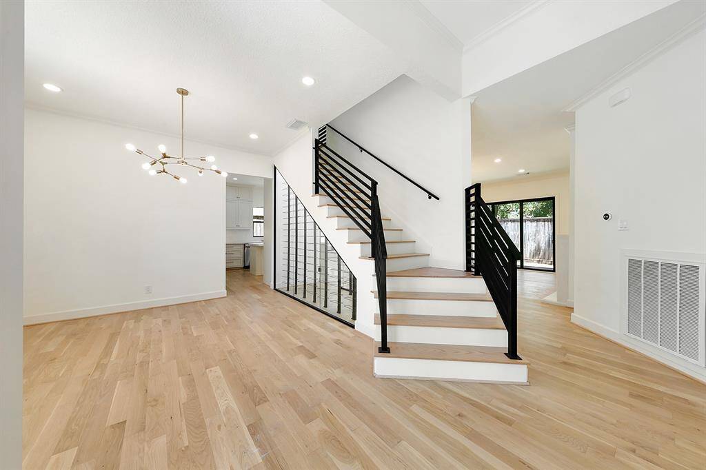 This photo showcases a modern, open-concept home interior with light wood flooring and a contemporary staircase featuring black railings. A stylish chandelier hangs in the dining area, and large windows provide natural light, creating a bright and inviting atmosphere.