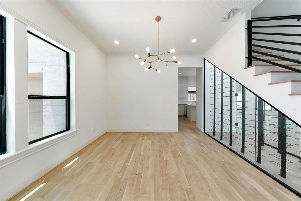 Bright and modern dining area with large windows, light wood flooring, a stylish chandelier, and a sleek glass railing along the staircase. The space leads into a kitchen area, offering an open and airy feel.