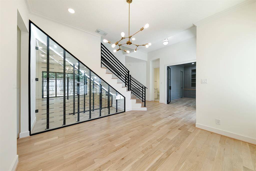 This modern entryway features a sleek staircase with glass paneling and black railing, complemented by light wood floors. A stylish chandelier adds elegance, while an open doorway leads to a home office or study area. The space is bright and airy with a contemporary design.