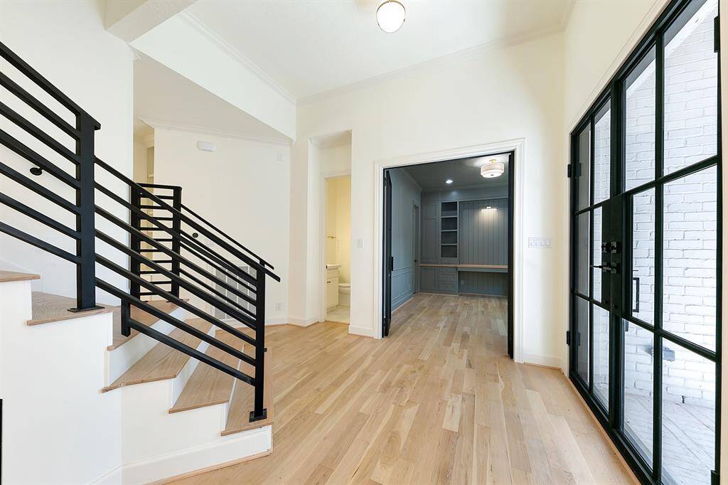 This photo showcases a modern entryway with light wood floors and a staircase featuring sleek black railings. It opens to a bright home office with built-in shelves and a desk. Large glass doors lead to an outdoor space, allowing plenty of natural light.
