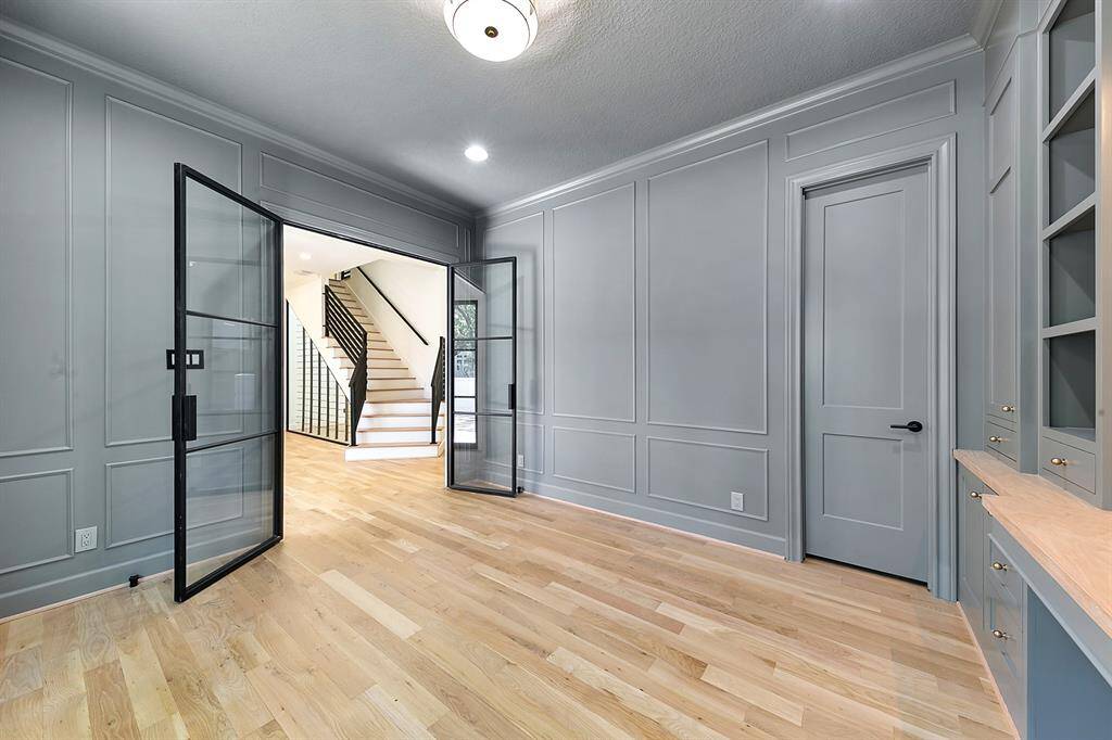 This room features elegant gray paneled walls, light hardwood flooring, and stylish glass double doors leading to a bright space with a staircase. Built-in shelving and cabinets offer functional storage.
