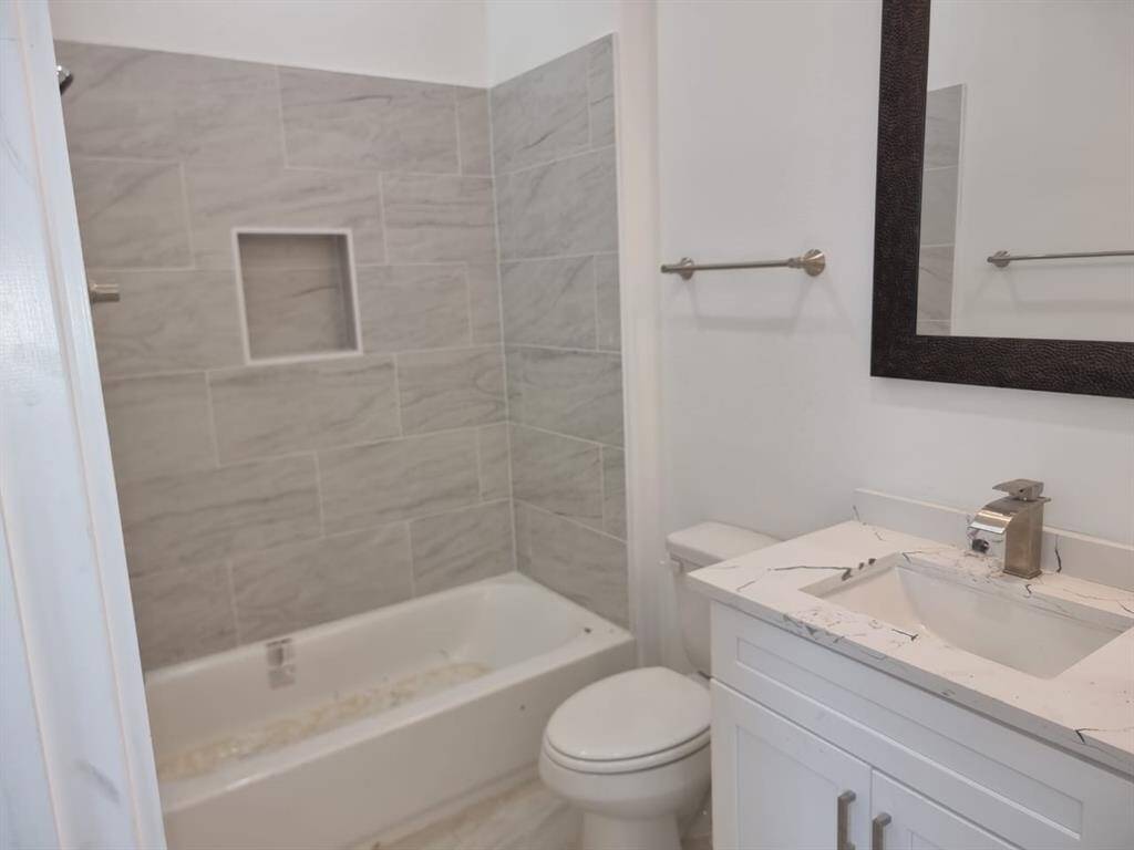 Modern bathroom featuring a bathtub with gray tile surround, a white vanity with a sleek faucet, and a large mirror. The space includes a toilet and towel bars, offering a clean and functional design.