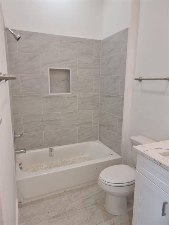 Bathroom features a modern design with light gray tile walls and flooring, a built-in bathtub, and a convenient shower niche. It includes a white vanity with a countertop and a standard toilet, offering a clean and functional space.