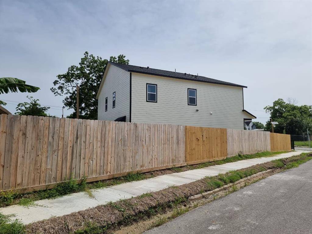 The photo shows a modern two-story house with light-colored siding and a dark roof, surrounded by a tall wooden fence. It is situated on a corner lot with a sidewalk and a few trees in the background, providing a sense of privacy and a suburban feel.