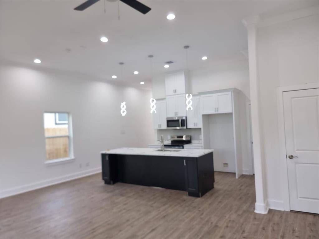 Modern open-concept kitchen with high ceilings, recessed lighting, and wood flooring. Features a central island with pendant lights, white cabinetry, and stainless steel appliances.