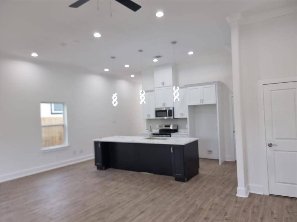 This photo showcases a modern, open-concept kitchen and living area with high ceilings, recessed lighting, and a ceiling fan. The kitchen features white cabinetry, stainless steel appliances, and a central island with pendant lighting. The space has a bright, airy feel with hardwood floors and a window providing natural light.