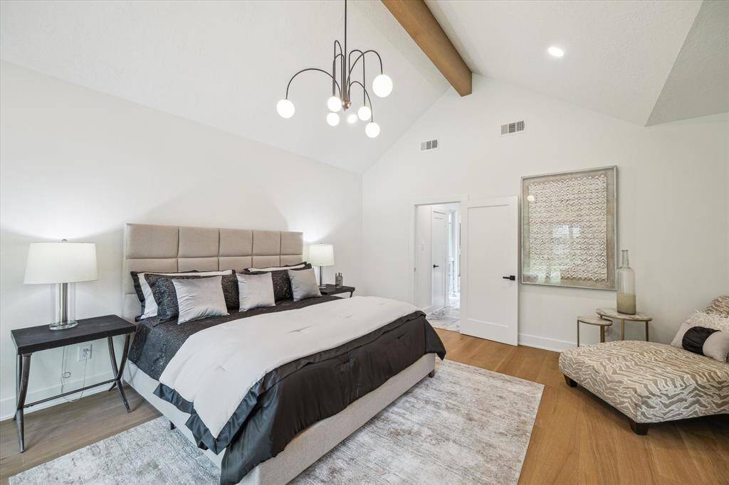 This spacious first floor primary bedroom features a high vaulted ceiling with exposed beams, a modern chandelier, and ample natural light. The room has a neutral color palette and connects to an en-suite bathroom.