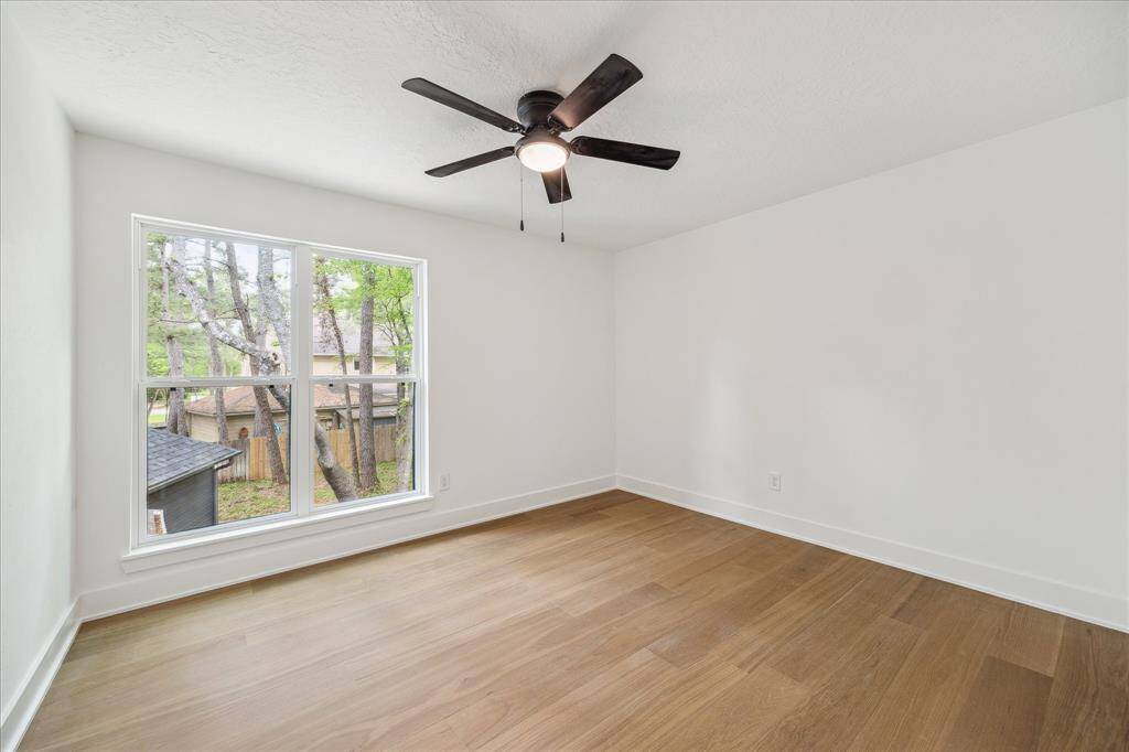 This bright room features large windows with a view of trees, light wood flooring, and a modern ceiling fan, creating an inviting and airy space.