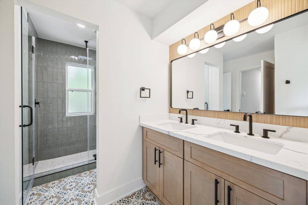 This modern bathroom features a spacious double vanity with sleek black fixtures, a large mirror with stylish lighting, and a glass-enclosed shower with green tiles. The patterned floor adds a touch of elegance.