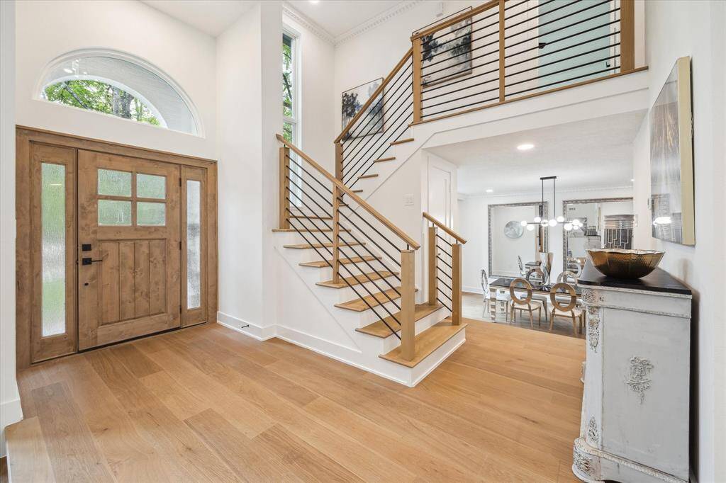 Bright and welcoming entryway featuring a rustic wooden front door, sleek staircase with modern railing, and an open view into a stylish dining area. The space is accented by light wood flooring and high ceilings, creating an airy, elegant atmosphere.