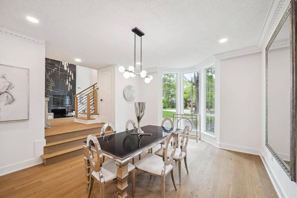 This elegant dining area features a modern chandelier, large bay windows for natural light, and hardwood floors. The space connects to a stylish stairway with a sleek railing, leading to another level. The room is bright and inviting, perfect for entertaining and special dinners.