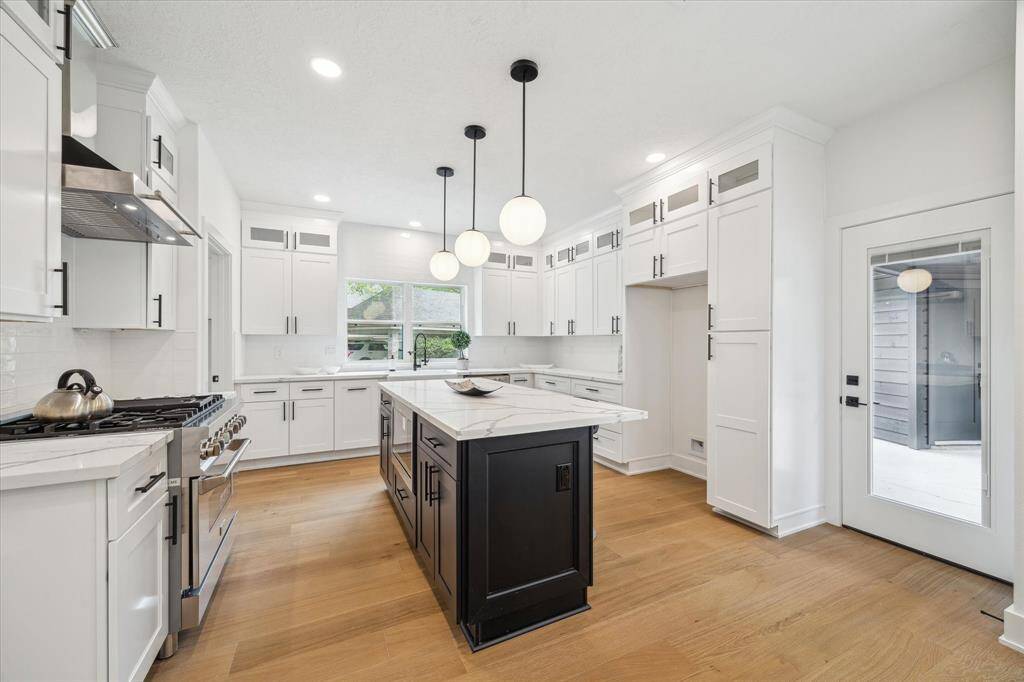 This modern kitchen features sleek white cabinetry, a large central island with a contrasting dark finish, and elegant pendant lighting. It includes stainless steel appliances, ample natural light from large windows, and light wood flooring, creating a bright and inviting space.
