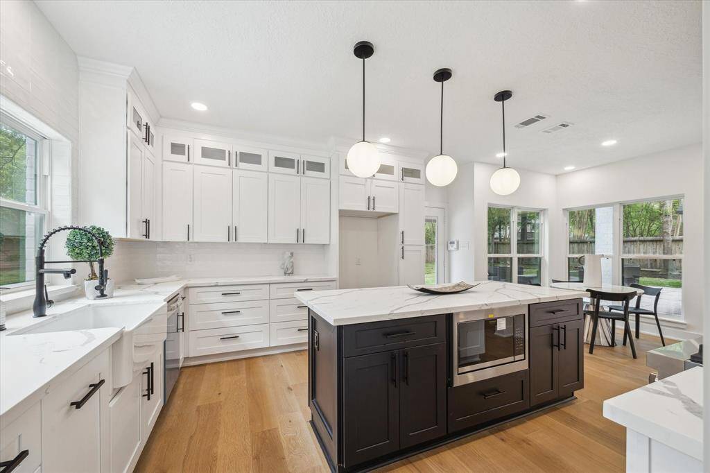 This kitchen features a modern, open-concept design with sleek glass front cabinetry, a large island with a built-in microwave, and elegant pendant lighting. Ample space with accessibility to the dining area overlooking the gardens.