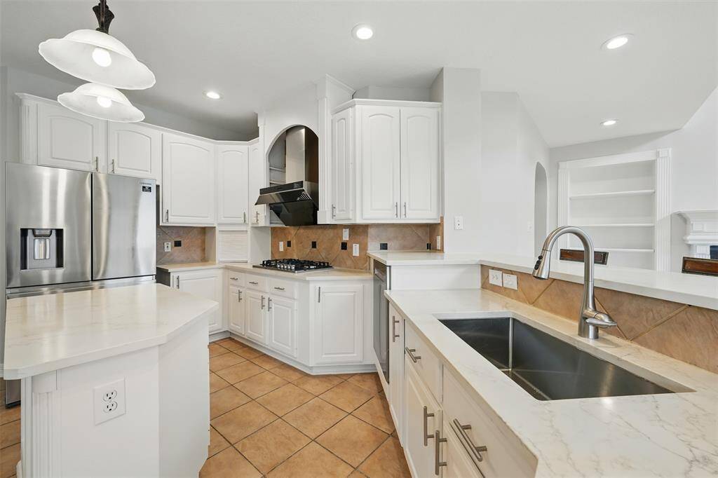 Bright, modern kitchen featuring white cabinetry, stainless steel appliances, and a spacious island. The room has a tiled floor, recessed lighting, and ample counter space with a double sink and elegant fixtures.