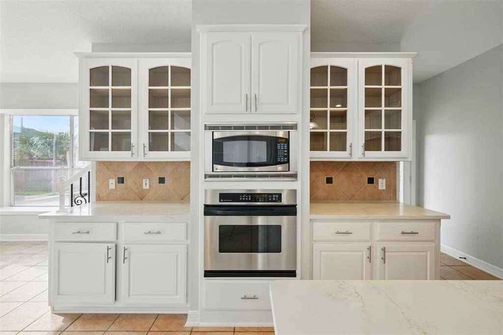 This kitchen features white cabinetry with glass-front doors, a built-in microwave and oven, and a tile backsplash. The space is bright, with ample natural light and tile flooring.