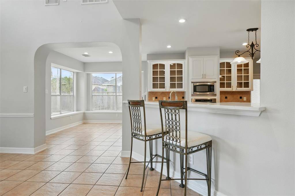 Bright and spacious kitchen with modern white cabinetry and open shelving, adjacent to a large breakfast nook with expansive windows. The area features tile flooring and a breakfast bar with seating.