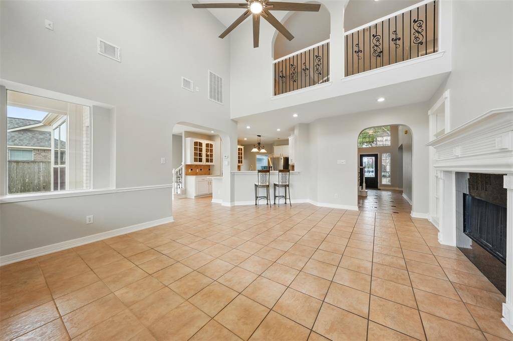 Spacious living area with high ceilings and tile flooring, featuring a fireplace and second-floor balcony. Open layout connects to a modern kitchen with a breakfast bar and ample natural light.