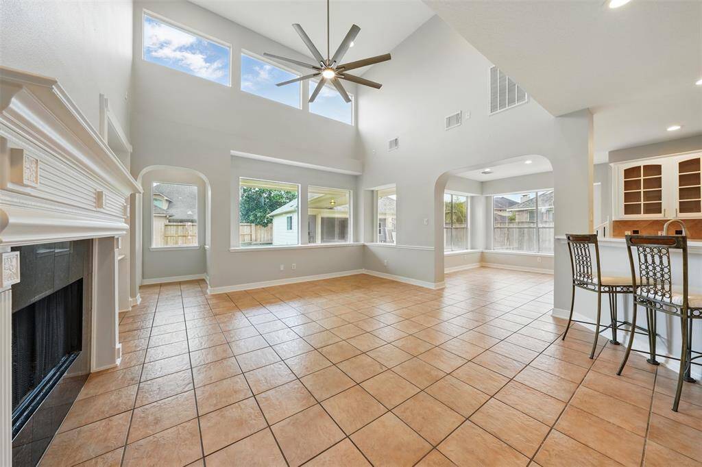 This bright and spacious living area features high ceilings, large windows, and a ceiling fan. The open concept design flows into a kitchen area with bar seating, and a cozy fireplace adds charm. Perfect for entertaining and family gatherings.