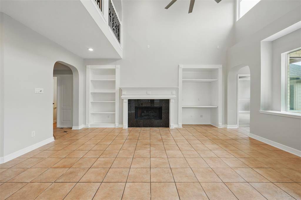This spacious family room features high ceilings, a cozy fireplace, built-in shelves, and large windows for natural light. The tiled floor enhances the room's elegance, and arched doorways add a touch of charm.