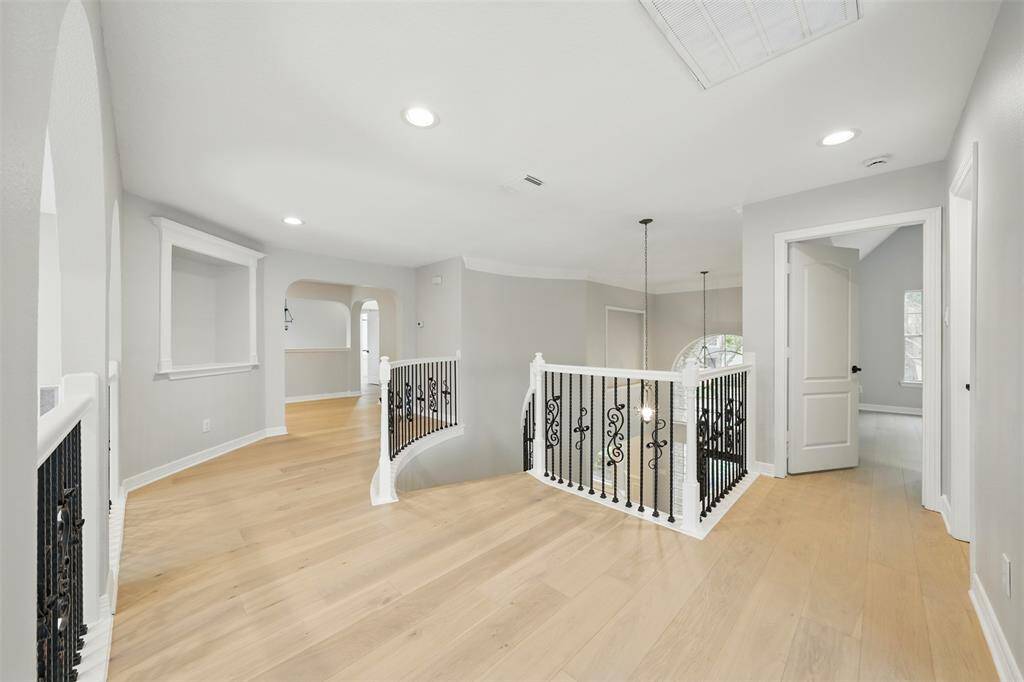 Open upstairs landing with light wood flooring and recessed lighting. It features elegant white and black wrought iron railings, multiple doorways leading to adjacent rooms, and an arched window providing natural light.