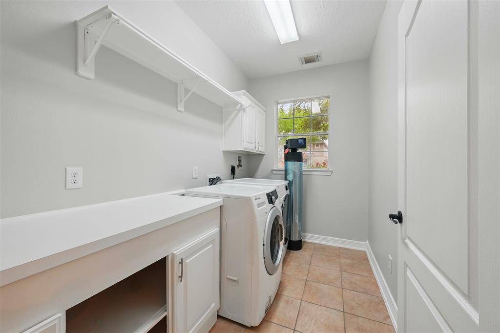Laundry room with a washer and dryer, white cabinetry, a countertop, and a shelf for storage. A window provides natural light, and the room has tiled flooring.
