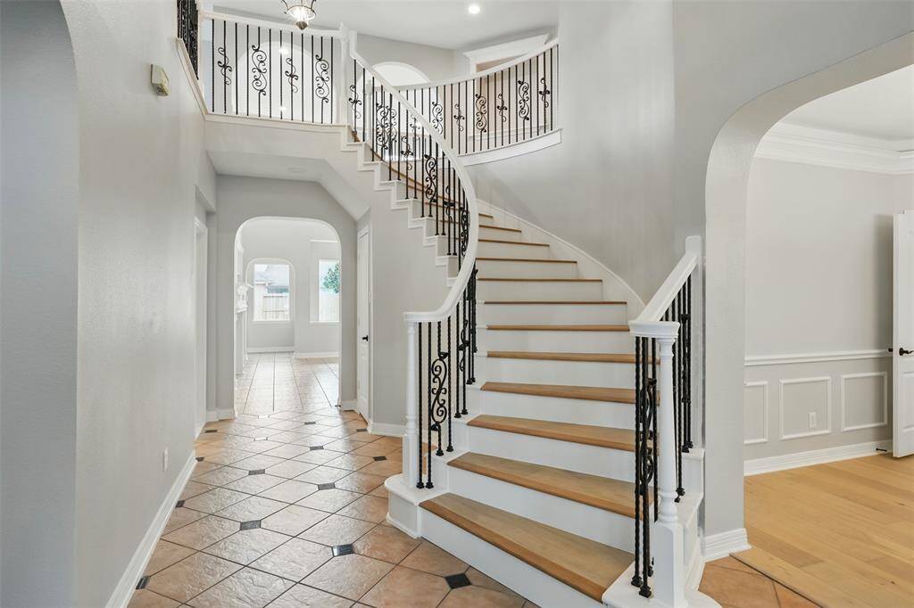 This elegant foyer features a grand, curved staircase with ornate iron railings, leading to an open, airy upstairs. The hallway is lined with stylish tile flooring and archways, offering a glimpse into a bright, spacious room. Ideal for welcoming guests with sophistication.