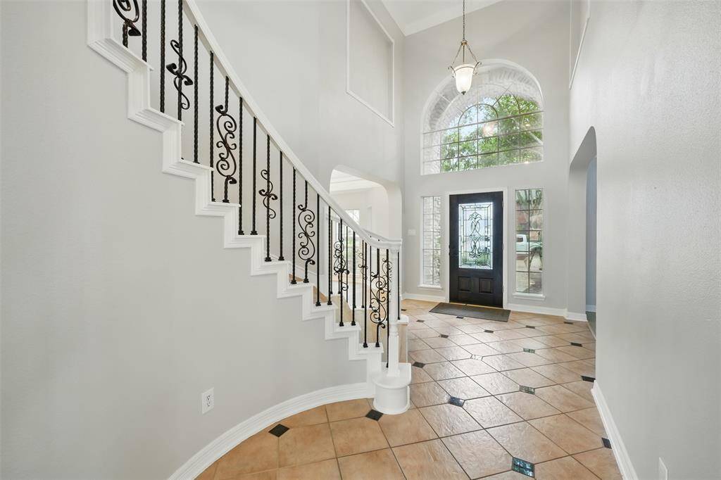 This photo showcases a bright, inviting foyer with high ceilings and large arched windows. The space features a stylish staircase with wrought iron railings, elegant tile flooring, and a decorative front door. Perfect for making a grand first impression.
