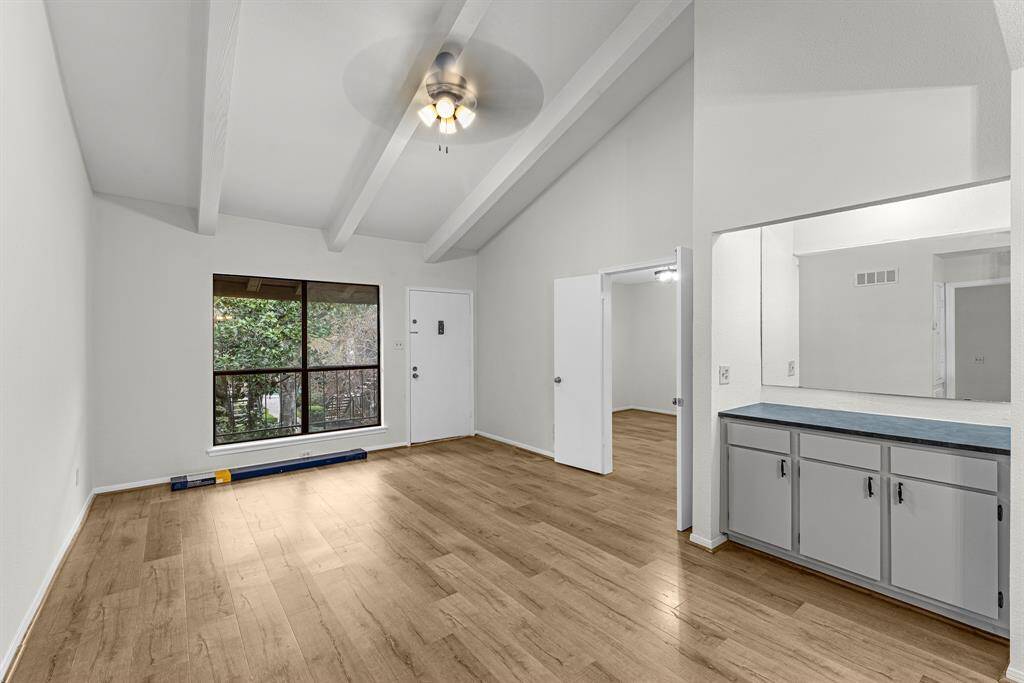 The living area features high ceilings with exposed beams, a ceiling fan, and large windows for natural light. The open layout with built in cabinet area, perfect for storage or a small bar. The laminate wood floors add warmth and elegance.