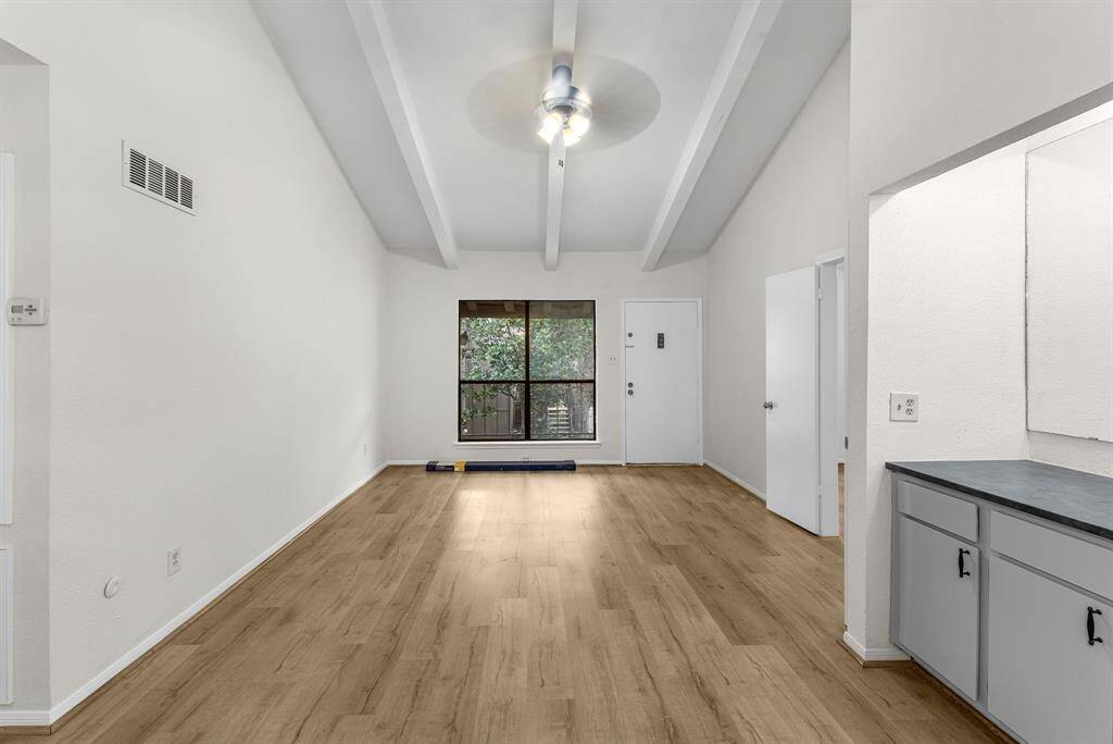 This room features a bright, open layout with high ceilings and exposed beams. The wood-look flooring adds warmth, and a large window provides natural light and a view of greenery. The space connects to a kitchen area with a countertop and cabinets, offering functionality and style.