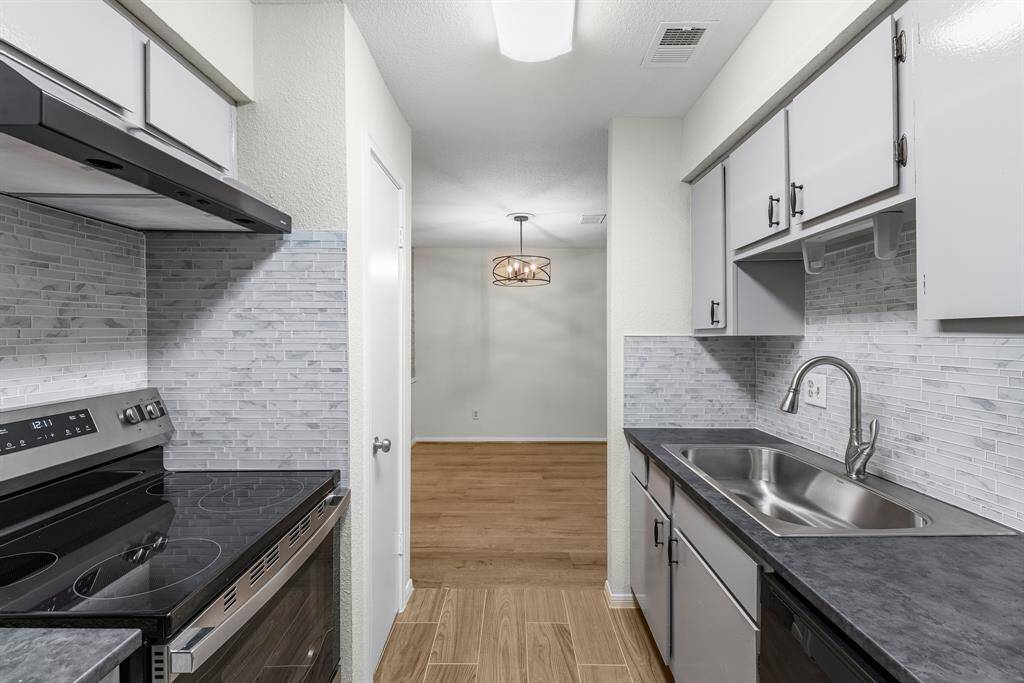 Modern kitchen with sleek grey cabinetry and countertops, featuring a stylish tile backsplash. Includes appliances and an oversized sink, with an open view to the dining area accented by a contemporary light fixture.