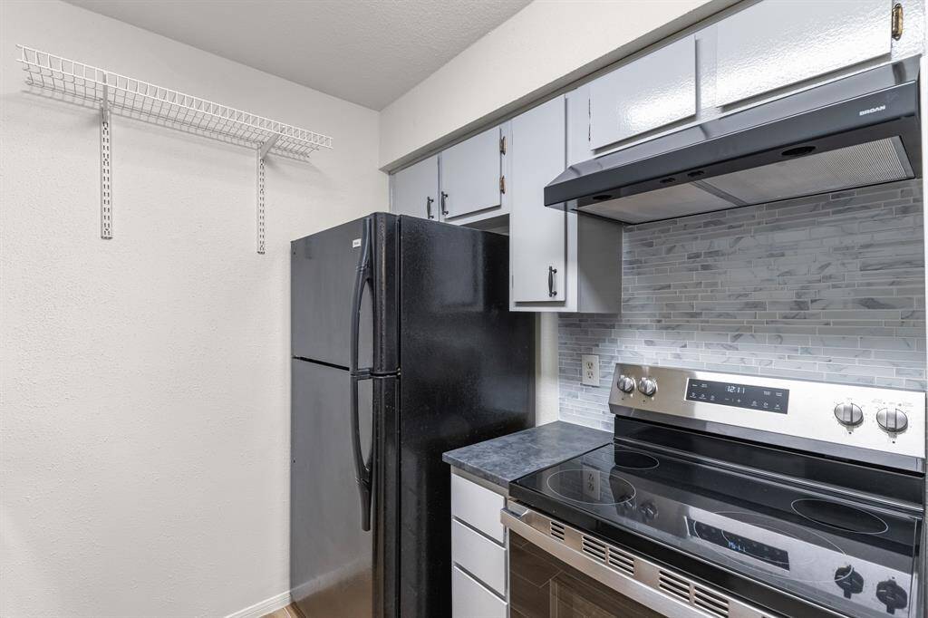 This kitchen features modern appliances, including a black refrigerator and a sleek electric stove with a stainless steel finish. It has updated cabinetry and a stylish tile backsplash, with additional shelving for storage.
