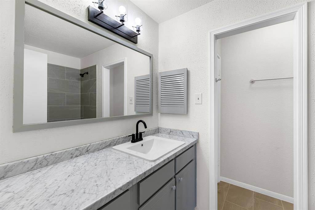 This bathroom features a modern design with a large mirror, marble countertop, and a sleek black faucet. It has a spacious vanity with storage and an adjacent walk-in closet. The neutral color palette and tile flooring add a clean, fresh feel.