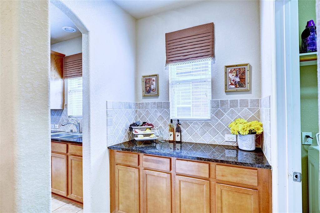 This photo showcases a cozy kitchen nook with warm-toned cabinets, granite countertops, and a tiled backsplash. It features a small window with a decorative shade, providing natural light. The space  has functional counter space when entertaining.