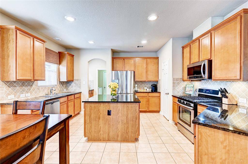 This kitchen features wooden cabinetry, stainless steel appliances, and a central island with a granite countertop. The space is well-lit with recessed lighting and has a tiled backsplash and floor, creating a warm and functional environment.