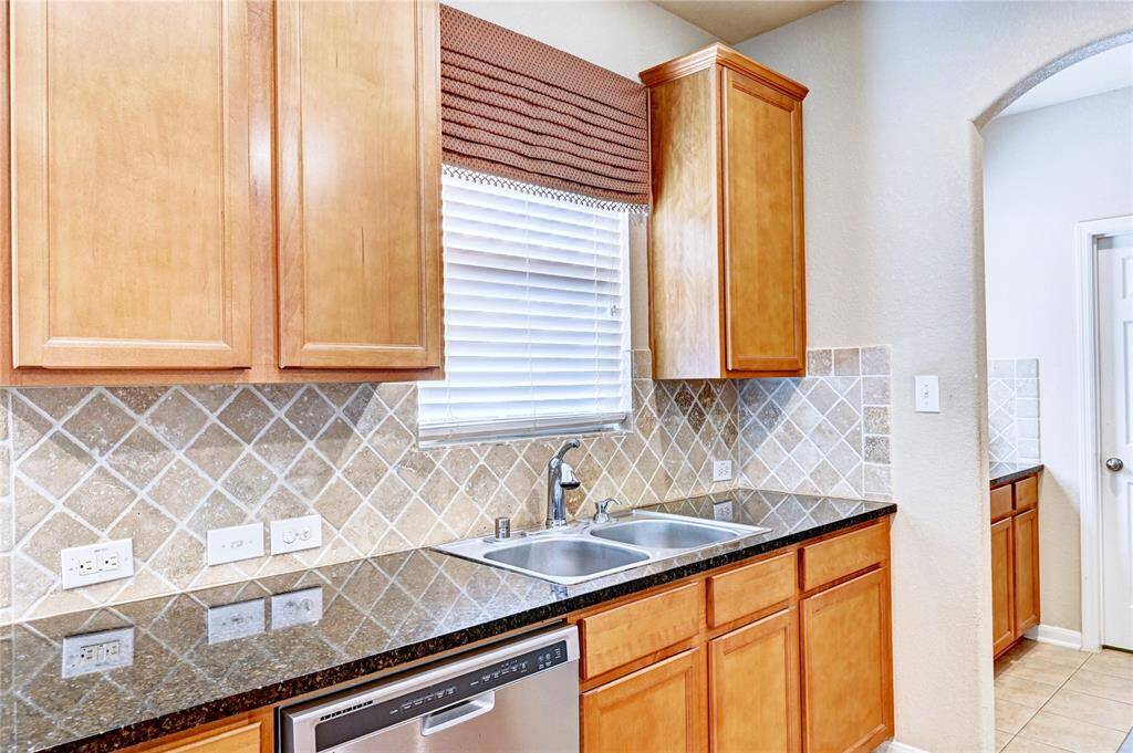 This kitchen features warm wood cabinets, granite countertops, a double sink, and a stainless steel dishwasher, complemented by a tiled backsplash and a window with blinds.