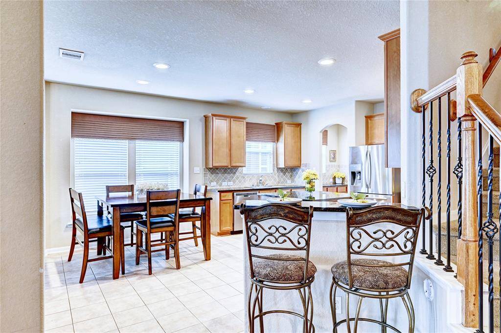 This photo showcases a bright and inviting kitchen and dining area. The space features modern stainless steel appliances, wooden cabinetry, and a cozy breakfast bar with ornate barstools. Adjacent is a dining table set near large windows, providing ample natural light. The open layout is complemented by tiled flooring and a staircase with decorative ironwork railing.