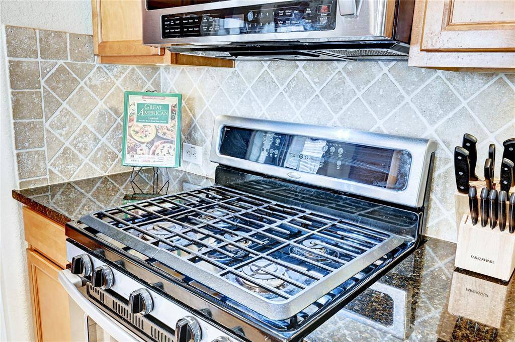 This kitchen features a modern gas stove with a stainless steel finish, set against a tiled backsplash. It includes a microwave above and granite countertops, offering a stylish and functional cooking space.