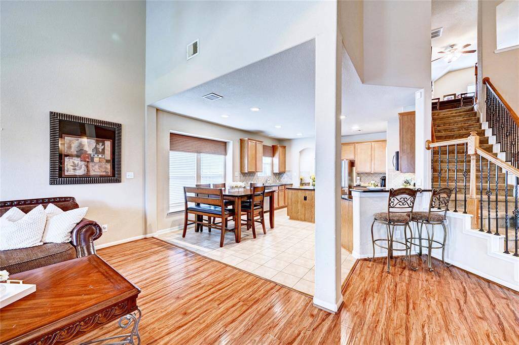 This photo showcases an open-plan living area with a cozy seating space, dining area, and a modern kitchen featuring wood cabinets and stainless steel appliances. The room has hardwood and tile flooring, plenty of natural light, and a staircase leading to an upper level.