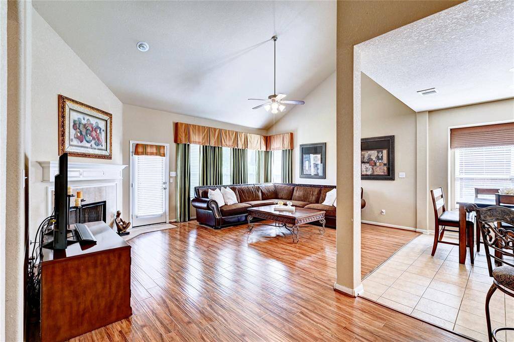 This photo showcases a bright, open living area featuring a cozy corner sectional, a fireplace, and hardwood floors. The living room flows into a dining area with tile flooring, enhancing the spacious feel. Large windows provide ample natural light, and a ceiling fan adds comfort.